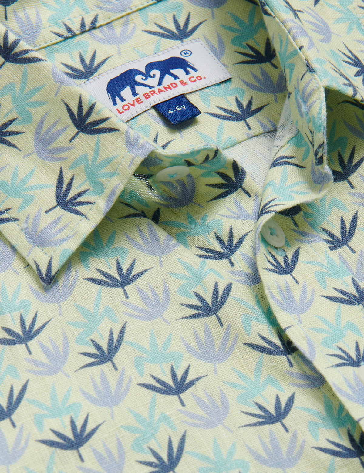 Close-up of the Boys Palm Sugar Abaco Linen Shirt featuring a tropical fan palm design in sky blue, aqua green, and deep blue on a limoncello yellow base, with a visible Love Brand & Co. label.