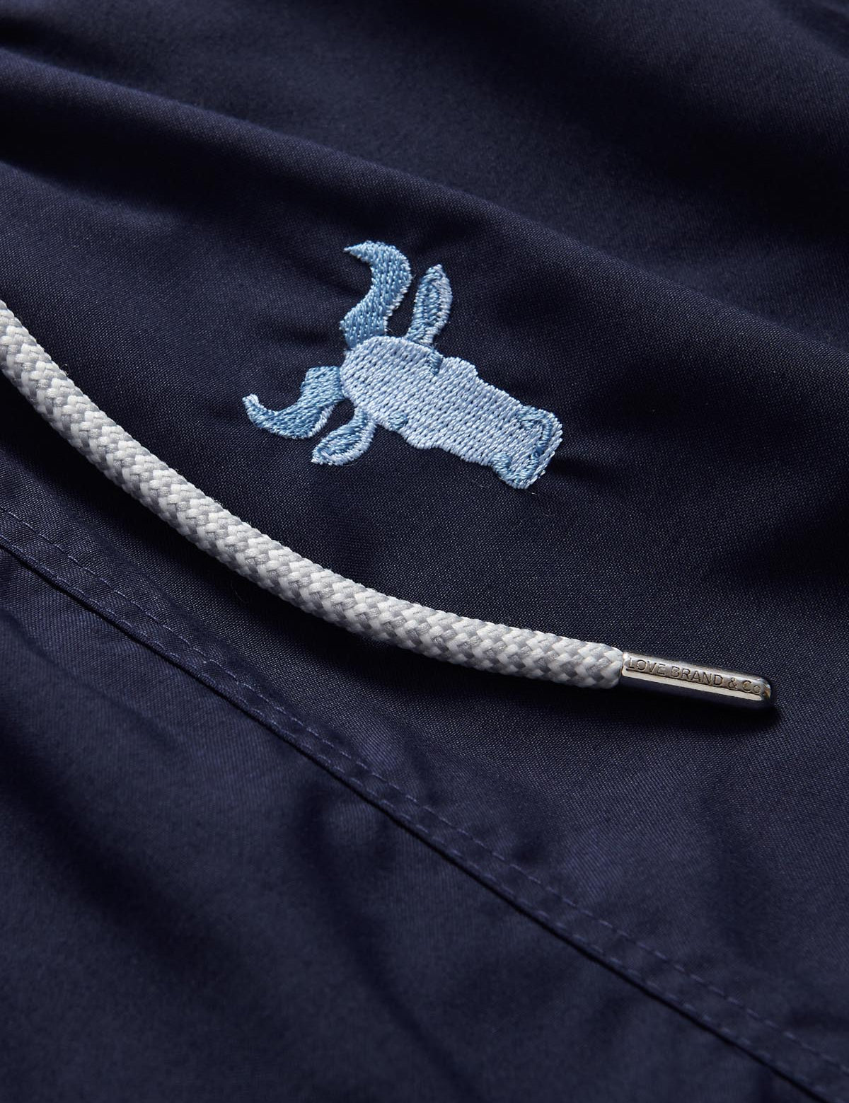 Navy blue men's swim shorts with intricate sky blue embroidered wildebeest design and white drawstring detail.