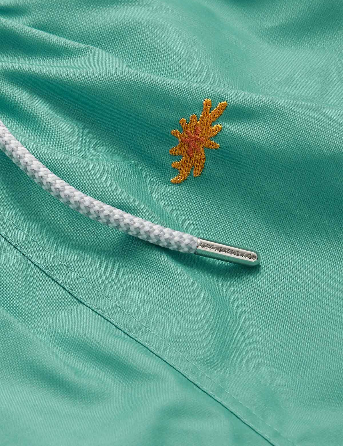 Close-up of the Men's Primavera Embroidered Staniel Swim Shorts in Riviera green. Embroidered swim shorts featuring an embroidered orange sea flower and drawstring detail.